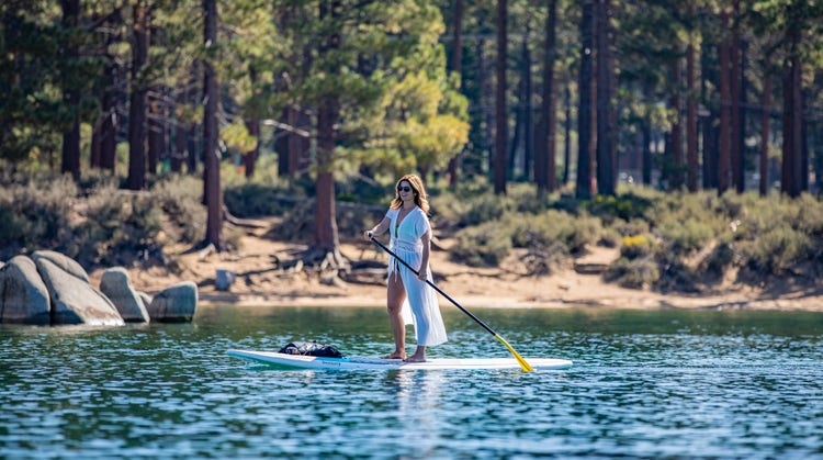 A woman stands on a paddleboard on a calm lake, holding a long paddle as she moves across the water.