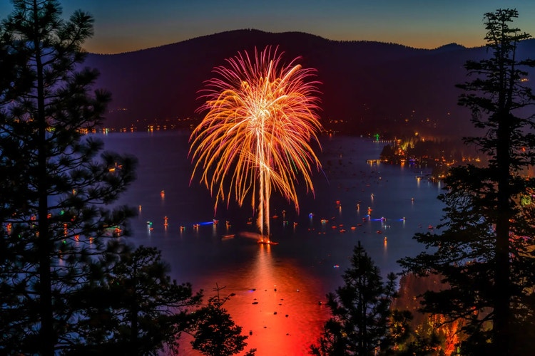 A large burst of colorful fireworks illuminates a calm lake at dusk, reflecting vibrant reds and yellows on the water