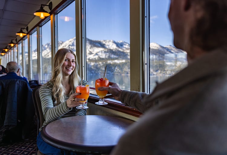 Couple seated at a table aboard a sightseeing cruise vessel, toasting colorful cocktails beside large windows with snow-covered mountains and a lake visible in the background.