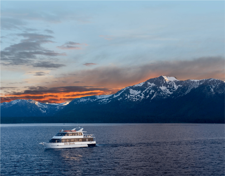 Tahoe Paradise yacht cruising across the calm waters of Lake Tahoe at sunset, with snow-capped Sierra Nevada mountains in the background and colorful orange and blue skies overhead.