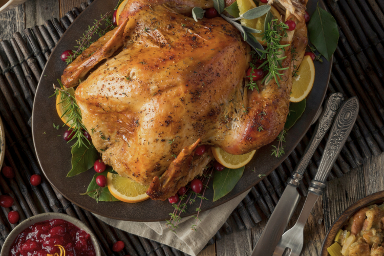 Roasted whole turkey garnished with fresh herbs, citrus slices, and cranberries on a serving platter, surrounded by traditional Thanksgiving side dishes including cranberry sauce and stuffing.