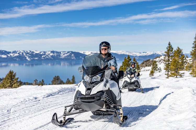 Two snowmobilers ride a snowy mountain trail overlooking Lake Tahoe and snow-covered peaks.